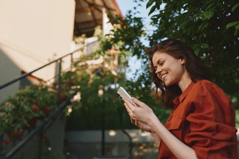 Young woman in a red blouse enjoys a sunny outdoor moment, smiling while checking her phone amid green leaves and warm daylight