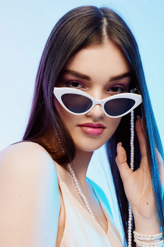 Young woman with long dark hair wearing white sunglasses and pearl jewelry poses against a blue background. Concept of beauty fashion and stylish accessories.