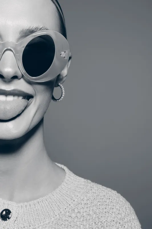 Young caucasian woman with light skin wearing large sunglasses and knitted sweater shows playful mood sticking out tongue.
