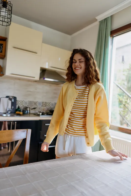 A cheerful woman in a yellow cardigan stands at a kitchen island, enjoying a relaxed morning at home. Bright daylight highlights casual lifestyle and domestic ease.