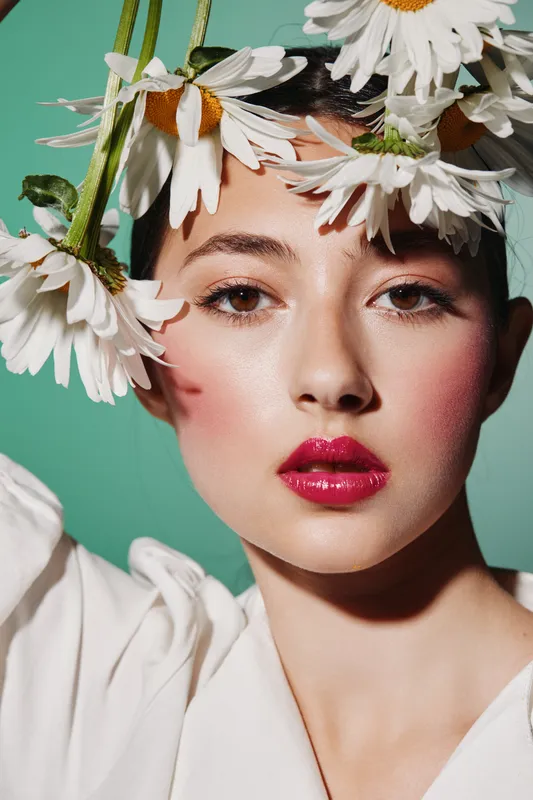 Beautiful woman with glowing skin and bright red lips poses with fresh daisies on a green background. Creative beauty and fashion portrait with natural floral decoration and soft lighting.
