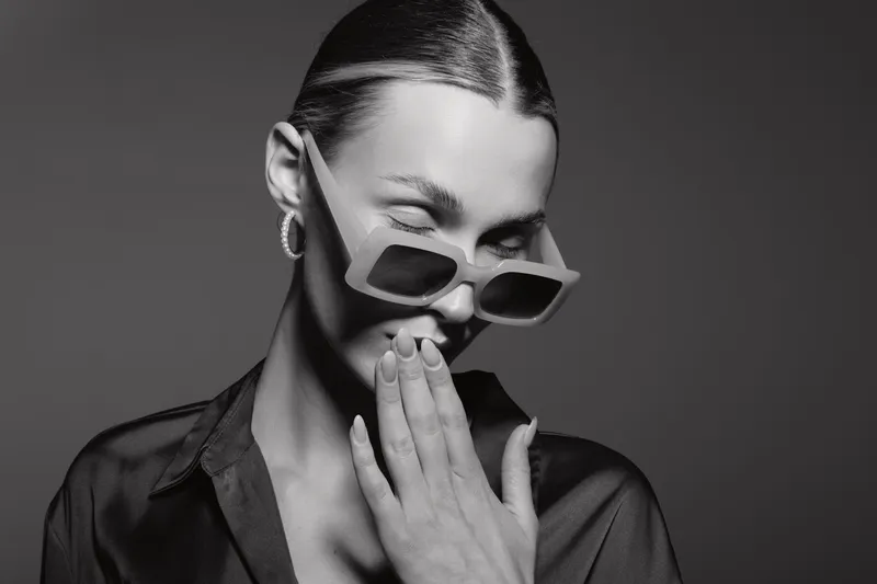 Young white woman with slicked hair wears sunglasses and smiles while covering mouth with hand in studio. Concept for fashion and emotion.