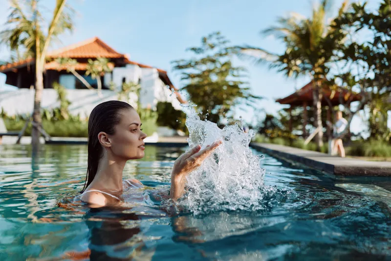 Young woman in swimming pool splashes water with hand enjoys sunny day in tropical resort with palm trees and villas in background lifestyle relaxation concept