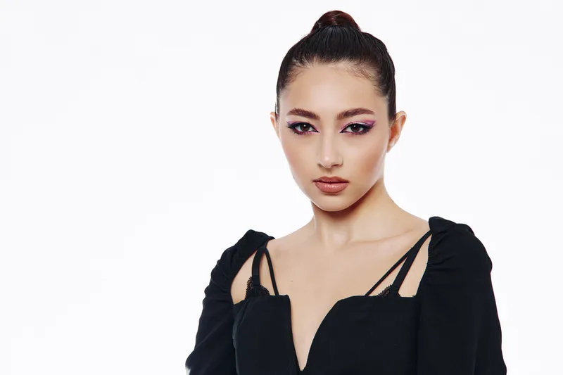 Young woman with sleek bun hairstyle wears black elegant top, looking seriously to camera. High contrast lighting with minimalistic white background. Fashion and beauty concept.