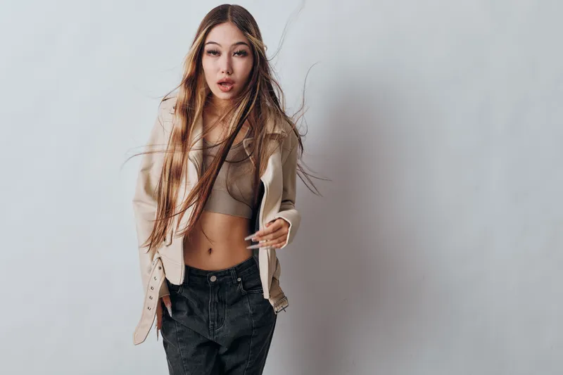 Young woman with long hair wearing open jacket and crop top posing confidently indoors with neutral background. Fashionable casual style and natural look with hands in pockets for portrait.