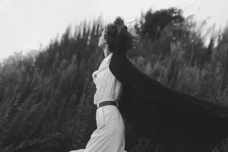 Cape, woman, wind, dress, outdoors, movement capture a dramatic figure as cape billows behind a flowing white dress in a windswept field, black and white photography emphasizing mood, freedom, and
