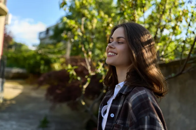Young woman enjoys a sunlit moment outdoors, smiling softly with a breeze in her hair, wearing a plaid shirt against a lush green backdrop