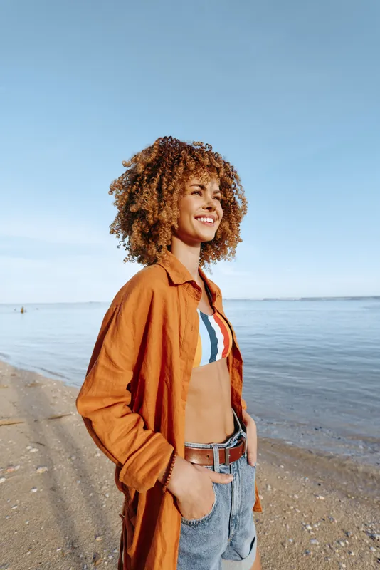 Happy young woman with curly hair standing on sandy beach wearing orange shirt and striped bikini top smiling with hands in pockets under clear blue sky by calm sea water