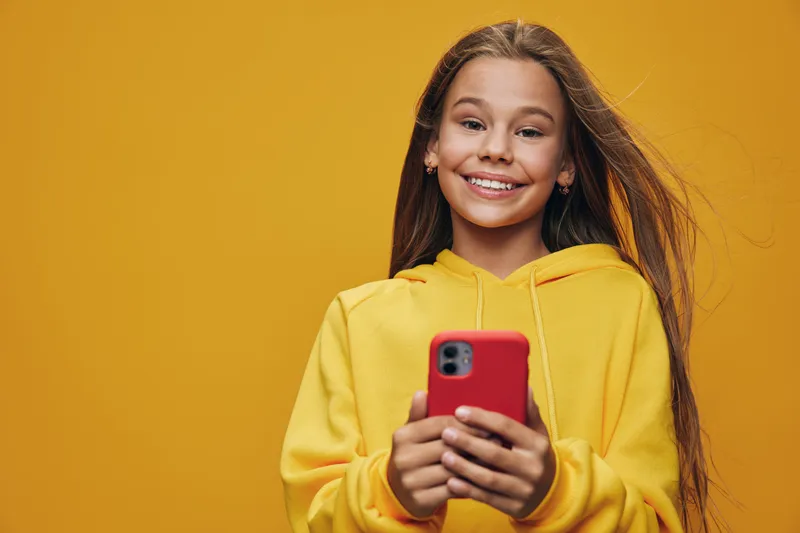 Girl smiling with long hair wearing yellow hoodie holding smartphone with both hands on yellow background. Happy child looking at camera with natural light and casual style indoors.