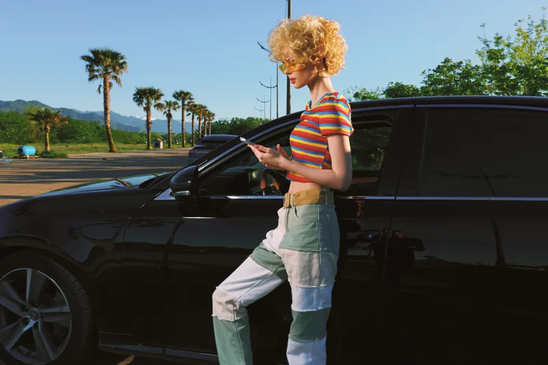 Woman lifestyle scene with retro fashion in outdoor setting by a car, striped top and jeans, warm film colors, sunny mood, casual pose, roadside vibe, bright and nostalgic.
