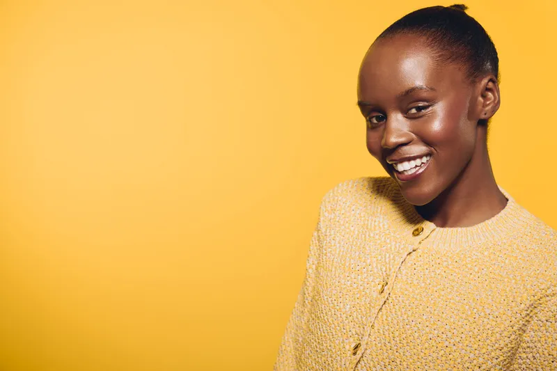 Smiling African American woman with dark skin wearing yellow sweater poses against bright yellow background. Concept of happiness, beauty, fashion, and positive mood with vibrant color.