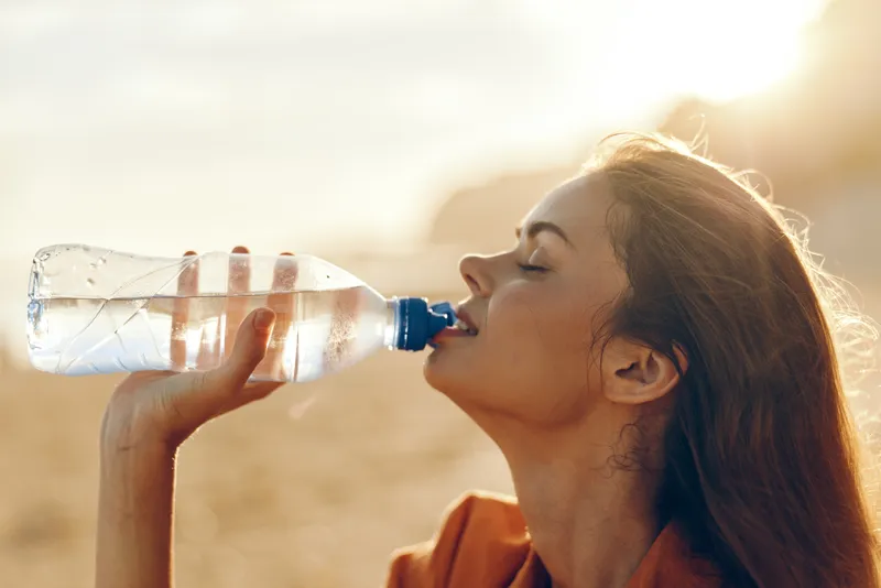 Woman drinking water from a bottle outdoors in bright sunlight close up profile view focus on hydration and thirst relief summer heat concept healthy lifestyle and refreshment after exercise in warm