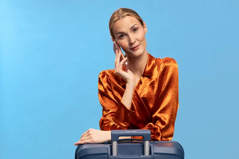 Young woman in shiny orange blouse poses with blue suitcase on blue background. Travel concept with confident female traveler and luggage in studio setting.