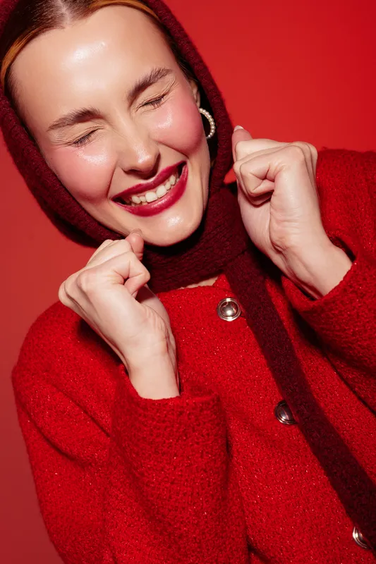 Happy woman in red sweater and scarf smiles with closed eyes and clenched fists on red background. Concept of joy, excitement, winter fashion, and positive emotions in studio lighting.