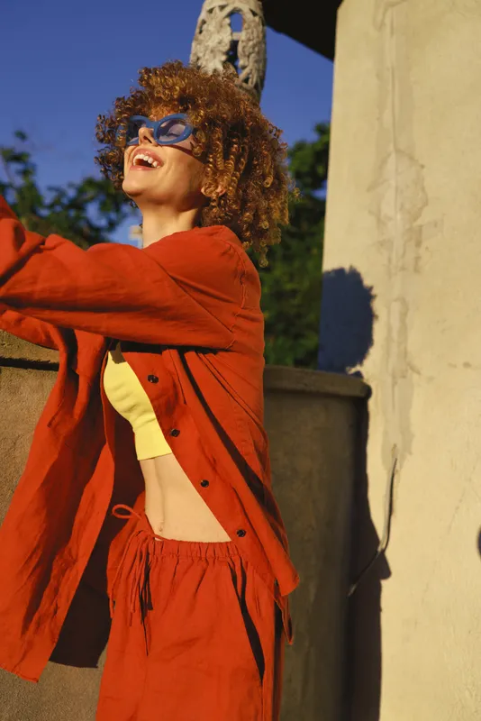 One woman with curly hair in an orange outfit smiles broadly outdoors, sunlight on a light wall, conveying confidence, energy, and carefree summer vibes as she moves freely