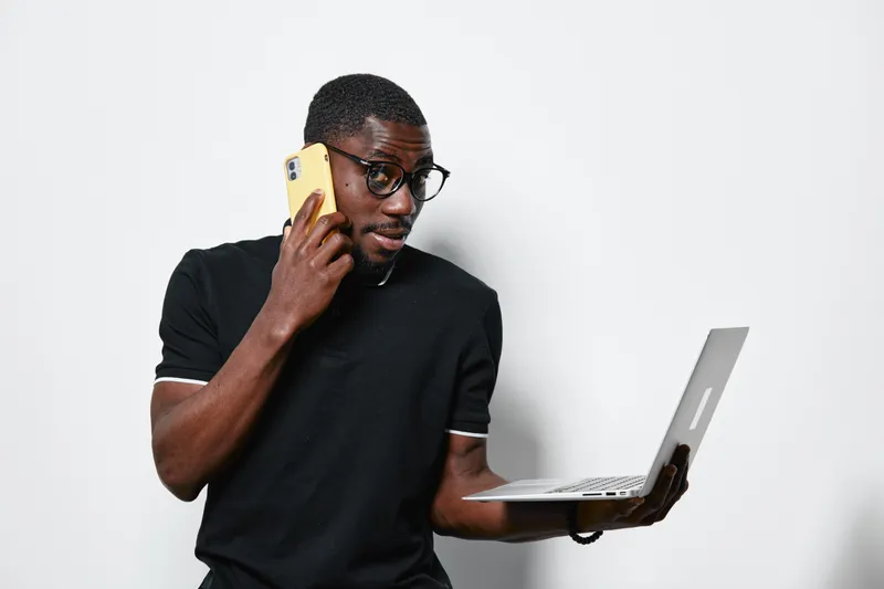 African American man with dark skin wearing glasses and black shirt talking on phone while holding laptop for remote work or communication in studio with white background.