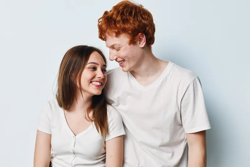 Smiling young couple with red hair and brunette looking at each other with happy expressions in casual white t-shirts on light background studio.