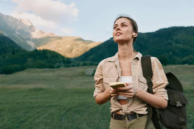 Young woman with backpack holding smartphone standing in green mountain landscape. Outdoor lifestyle, travel concept with natural light and peaceful mood.