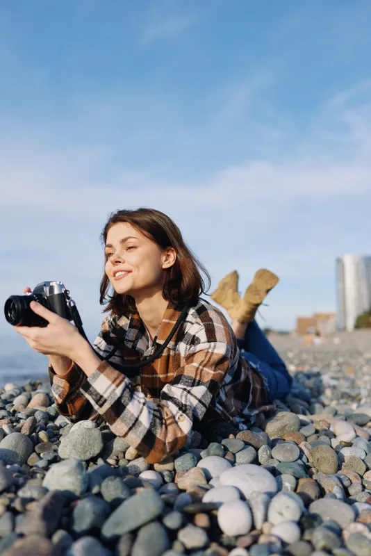 Woman lying on a pebble beach, holding a camera and smiling, casual plaid shirt, sunny day, blue sky, relaxed outdoor photography moment on the coast, capturing spontaneous scenes and natural light.