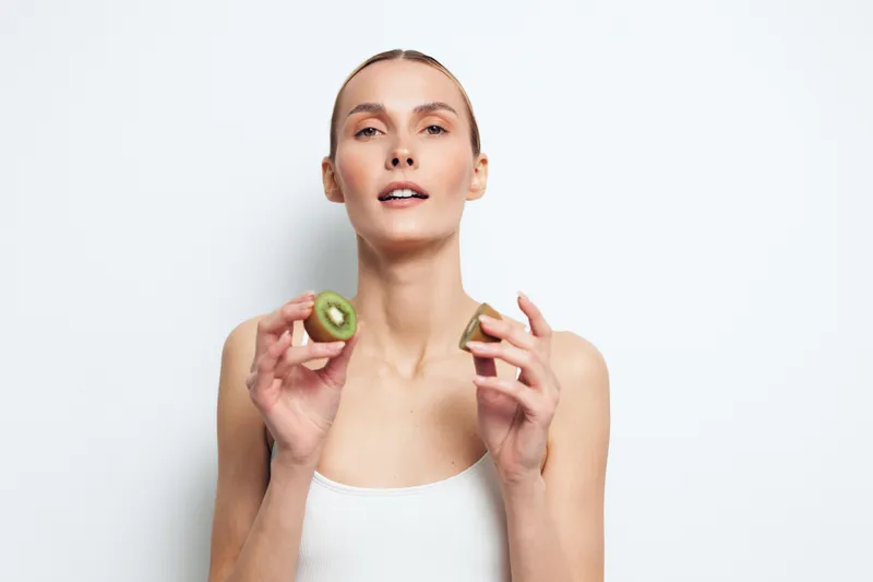 A healthy young woman in a white tank top holds two halves of fresh kiwi fruit near her face, symbolizing natural beauty, nutrition, and wellness on a clean white background.