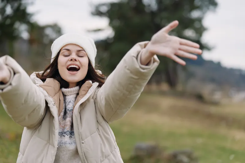Joyful young woman outdoors with arms wide open, wearing a puffy coat and knit hat, expressing freedom, happiness and carefree energy in a park on a chilly day.