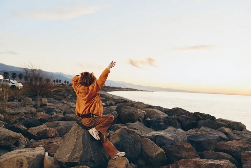 Woman lifestyle, rocky beach at sunset, ocean waves behind her, she sits on rocks with arms raised, relaxed portrait, freedom feeling in warm evening light, slow travel moment by the shore