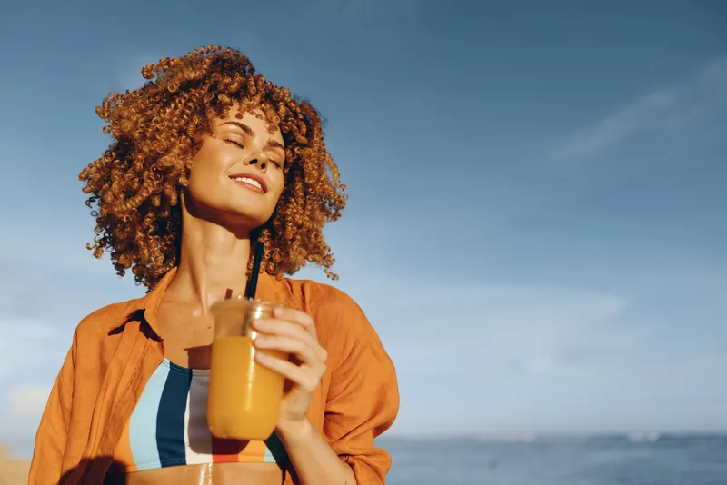 Smiling woman with curly hair holding a glass of orange juice outdoors near the sea under clear blue sky. Concept of healthy lifestyle, relaxation, and summer vacation.