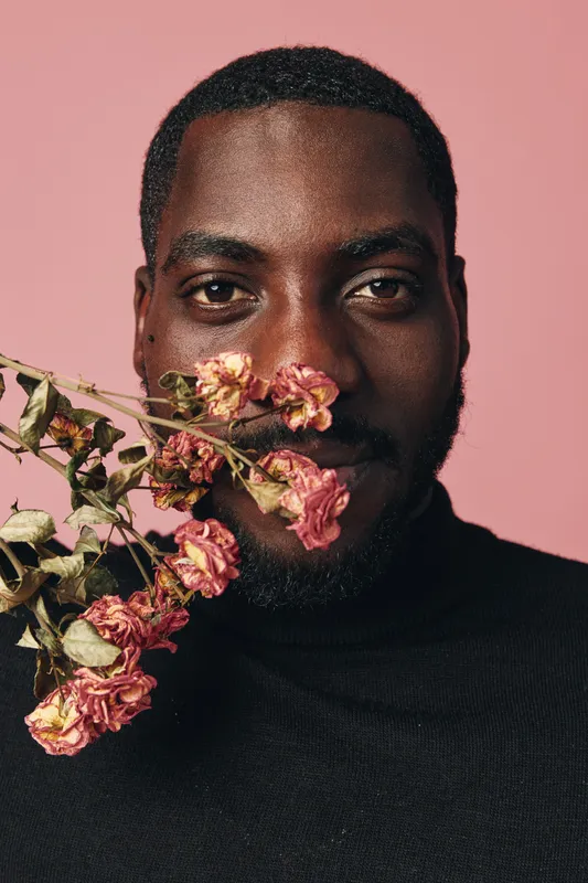 African man with dark skin and beard holding dried pink flowers near face in creative conceptual visual style with pink background. Portrait with artistic expression and natural texture.