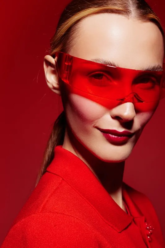Close-up portrait of confident woman wearing red transparent glasses and red outfit against red background. Concept of fashion, style, modern accessories, and bold makeup look.