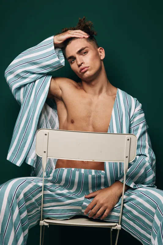 Young man with curly hair wearing striped pajamas sitting on chair backwards with hand on forehead against green background. Casual relaxed pose and thoughtful expression.
