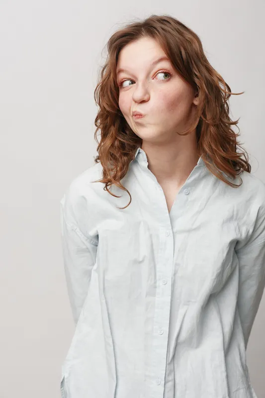Young woman with curly hair wearing white shirt looks sideways with a playful skeptical expression. Isolated on light background. Concept of doubt, curiosity, and casual portrait.