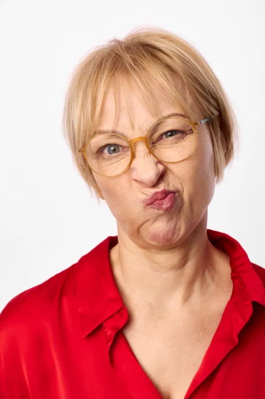Woman senior glasses portrait blonde studio expression red top on white background. Close up headshot of older woman with short hair and eyeglasses, soft studio lighting, calm friendly mood.