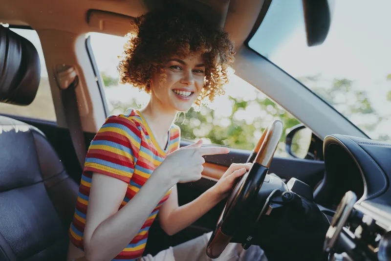 Cheerful woman driving, sunlit car interior, striped tee, candid lifestyle moment, gentle breeze, natural colors, relaxed smile, urban road visible through window, warm retro vibe, soft shadows