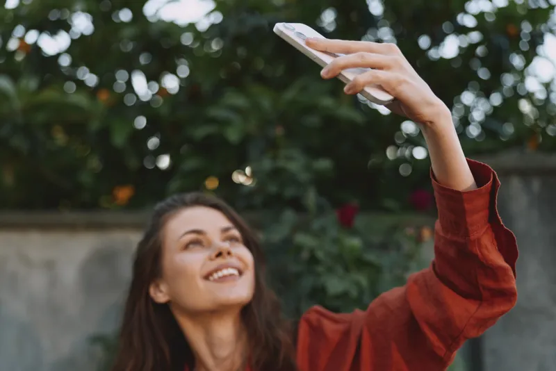 Smiling woman with long hair wearing a red sweater taking a selfie outdoors in a garden, showcasing casual lifestyle and self expression