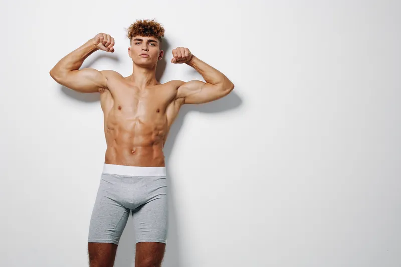 Young muscular man posing shirtless in gray shorts flexing biceps against white wall for fitness, bodybuilding, strength and healthy lifestyle concept with confident expression.