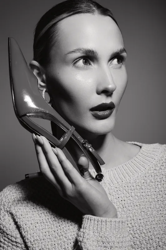 Elegant white woman with light skin holds shiny high heel shoe near face, wearing knitted sweater. Studio portrait in black and white with vintage fashion and beauty concept.