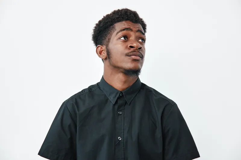 Young black man with dark skin wearing black shirt looking up thoughtfully on white background. Portrait of African ethnicity male with short curly hair and casual style.