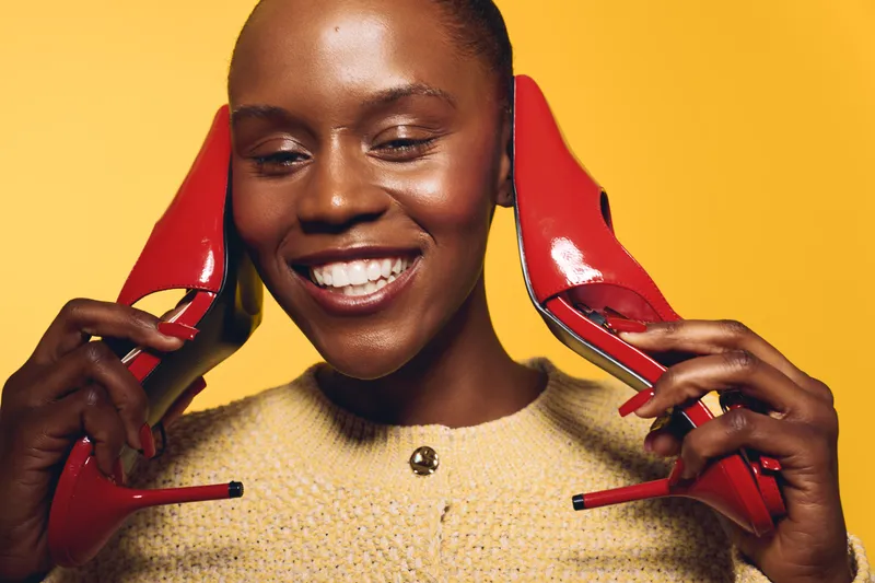 Smiling dark skinned African woman holds red high heels near her face against yellow background. Concept of fashion, beauty, happiness, and stylish footwear for women.