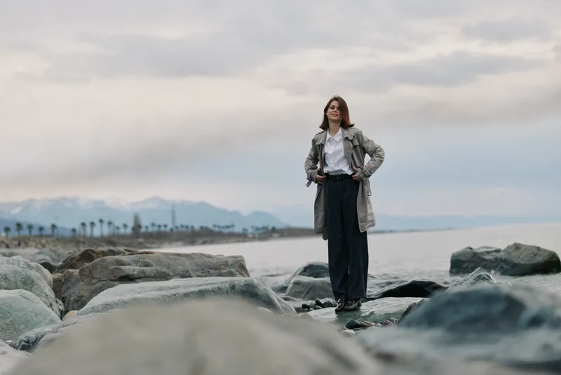 Confident woman in trench coat stands on rocky coastline with cloudy sky and distant mountains. Concept of outdoor fashion, freedom, and nature exploration with casual style.
