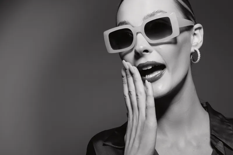 Stylish Caucasian woman with fair skin wearing large sunglasses and hoop earrings gestures with hand near mouth in black and white portrait.