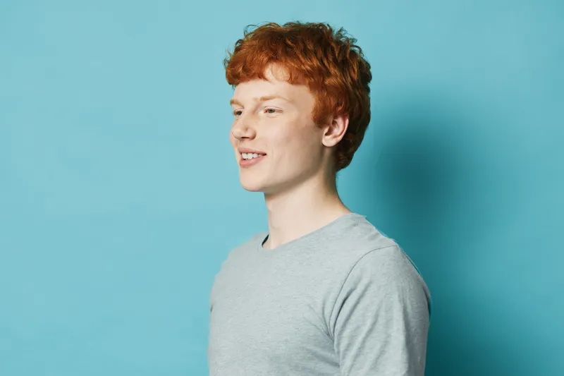 Young man with red hair and freckles smiling, wearing gray shirt standing against blue background in studio. Portrait of happy male model with casual style and natural expression.