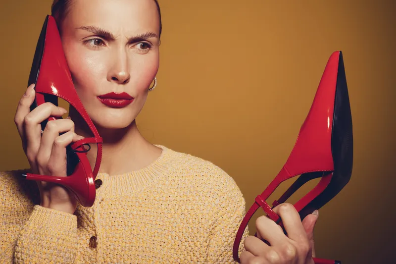 Woman in beige sweater holds red high heels near face with confused expression on yellow background. Concept of fashion dilemma, style choice, and footwear decision.