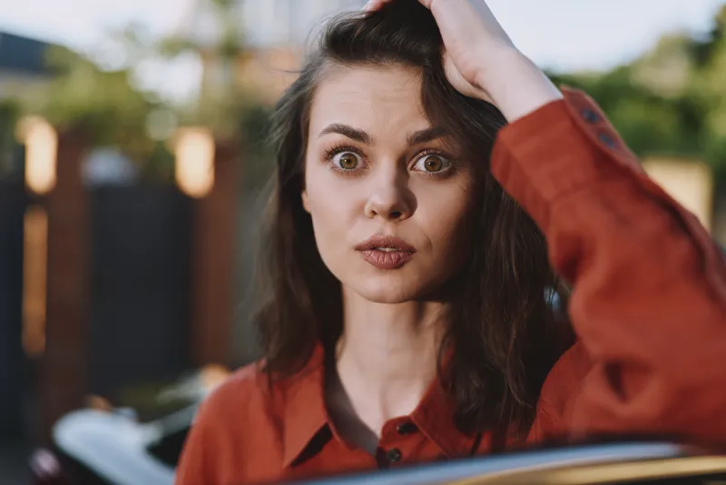 A surprised young woman with brown hair wears an orange shirt outdoors, looking at the camera with wide eyes, conveying curiosity, spontaneity, and a bold mood
