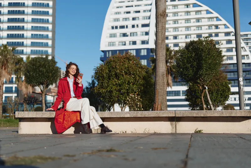 A stylish young woman sits on a bench in a sunny urban park, wearing a red coat and white pants, smiling softly with modern buildings in the background.