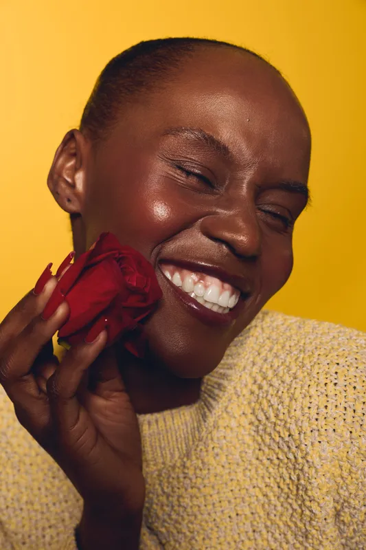 Close-up of happy dark skinned African woman smiling with closed eyes holding red rose near face. Concept of joy, beauty, natural skin, love, and positive emotions on yellow background.