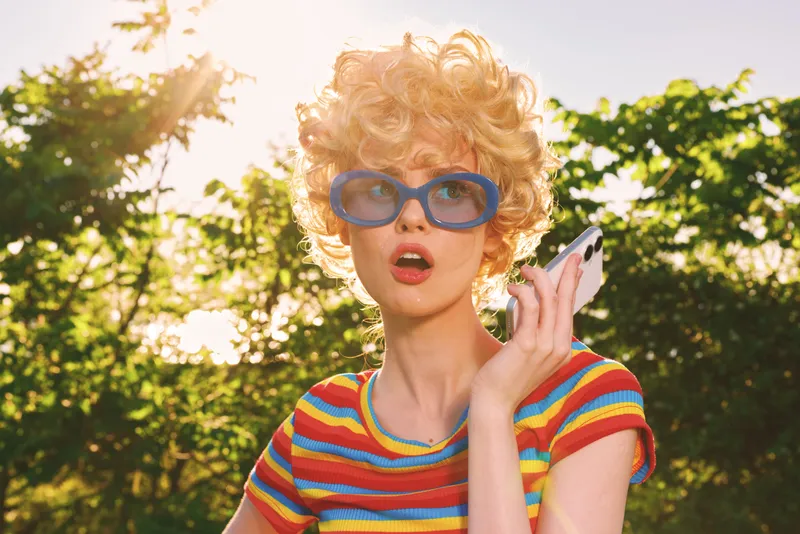 Woman outdoor retro lifestyle, wearing colorful stripes and sunglasses, talking on a phone, sunlit moment with breeze, vibrant color and nostalgic film mood