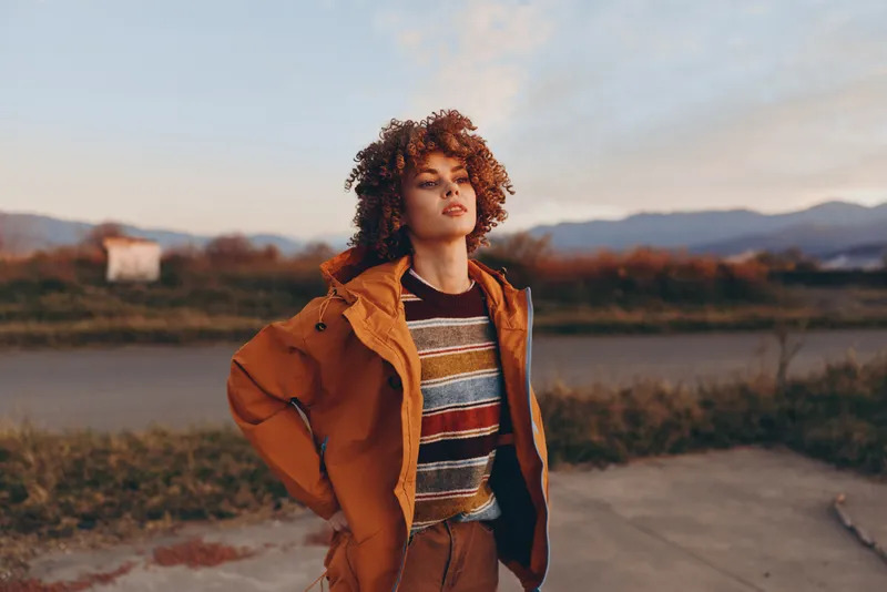 Woman lifestyle outdoors nature sunset jacket curly hair, river path countryside background, wearing brown jacket and striped shirt, relaxed walking pose, warm golden light