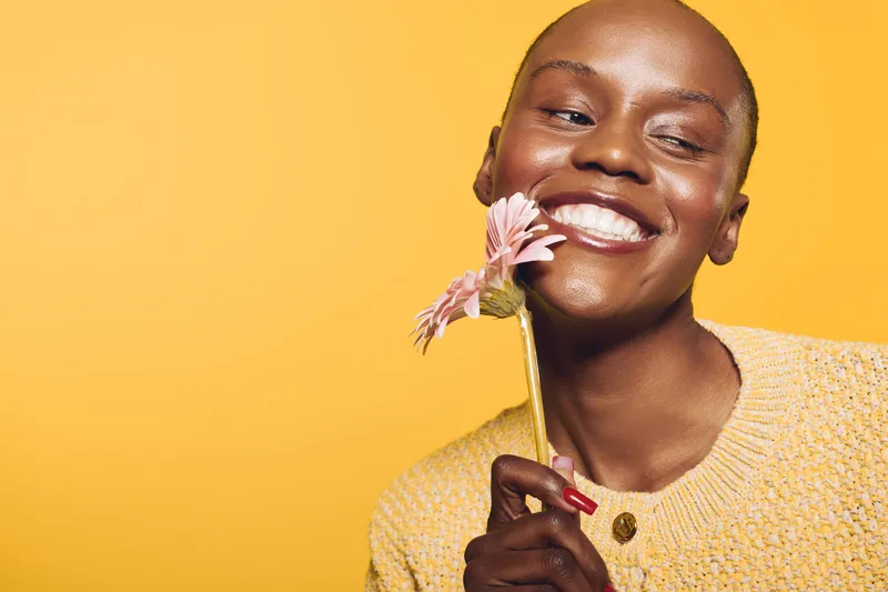 Happy African American woman with dark skin smiles holding pink flower near face on yellow background. Concept of joy, beauty, natural makeup, wellness, and positive mood in studio.