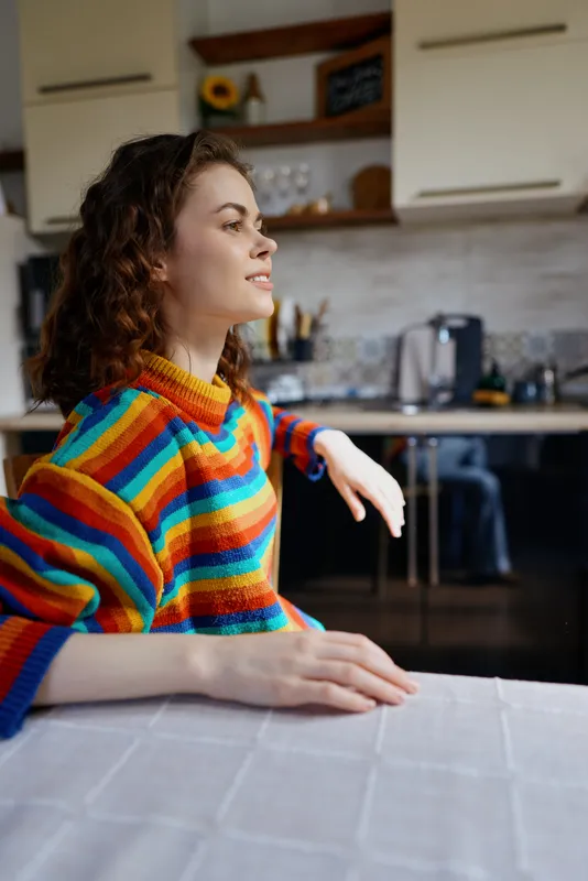 A woman in a striped colorful sweater sits at a table in a bright kitchen, leaning on her forearms, gazing to the side with a calm, thoughtful expression. Concept for relaxed home life and personal