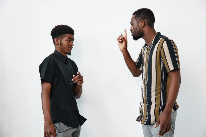 Two African American men with dark skin having serious conversation in casual clothes on white background. One man gestures while the other listens attentively in studio.